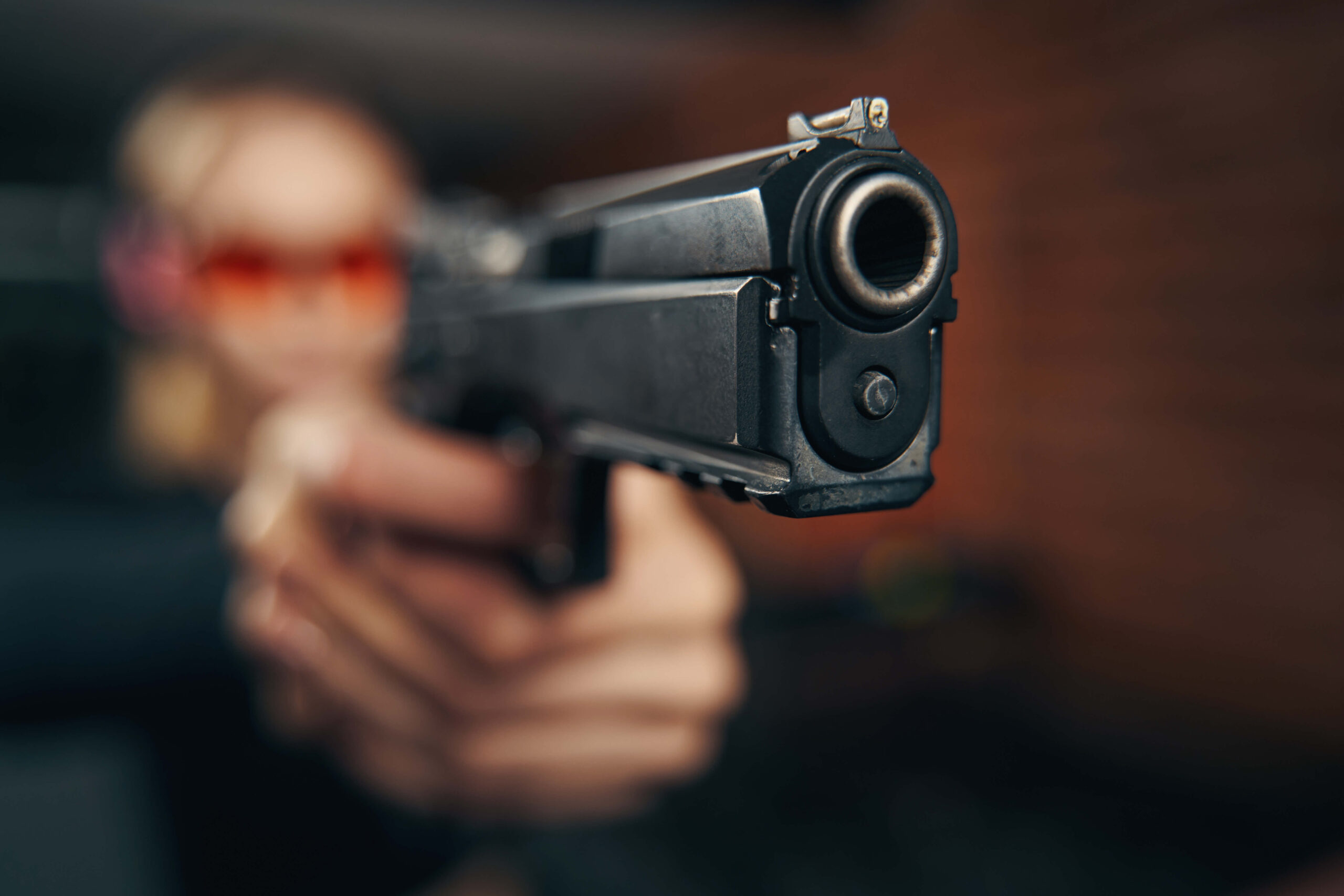 A close-up of a person aiming a handgun, focusing on the barrel and front sight. The person’s face is blurred in the background.