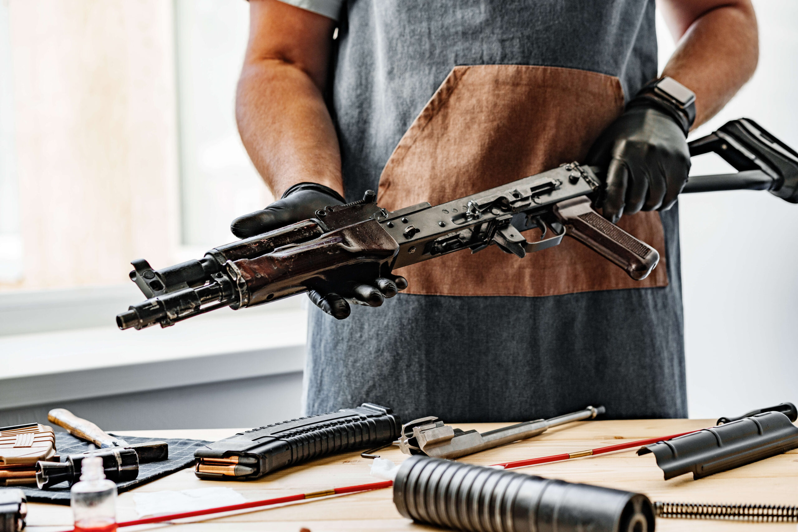 A person wearing gloves and an apron inspects a rifle. Various firearm components and tools are laid out on the table.
