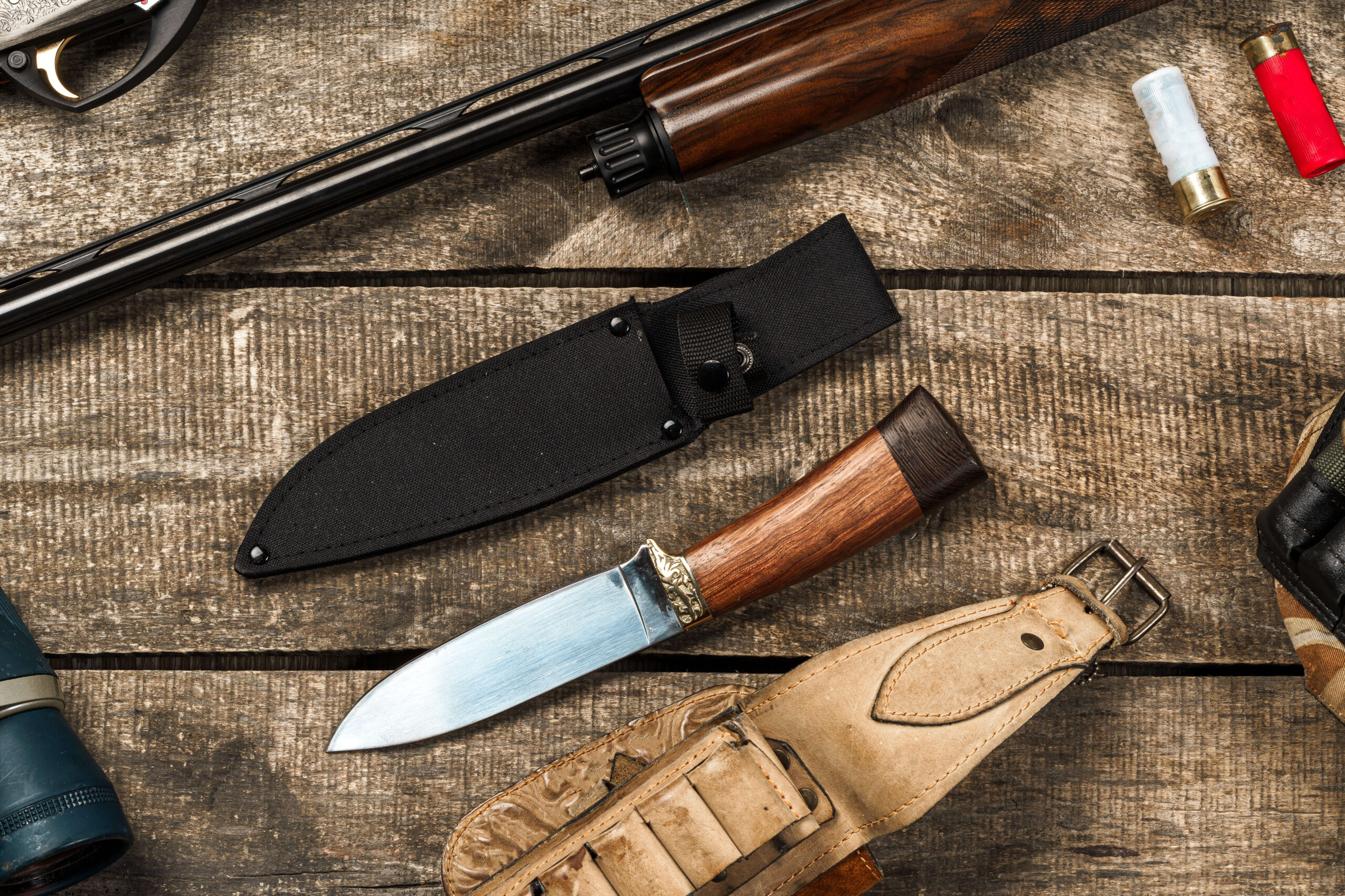A hunting knife with a wooden handle in a sheath, positioned beside a rifle, shotgun shells, and other hunting gear on a wooden surface.