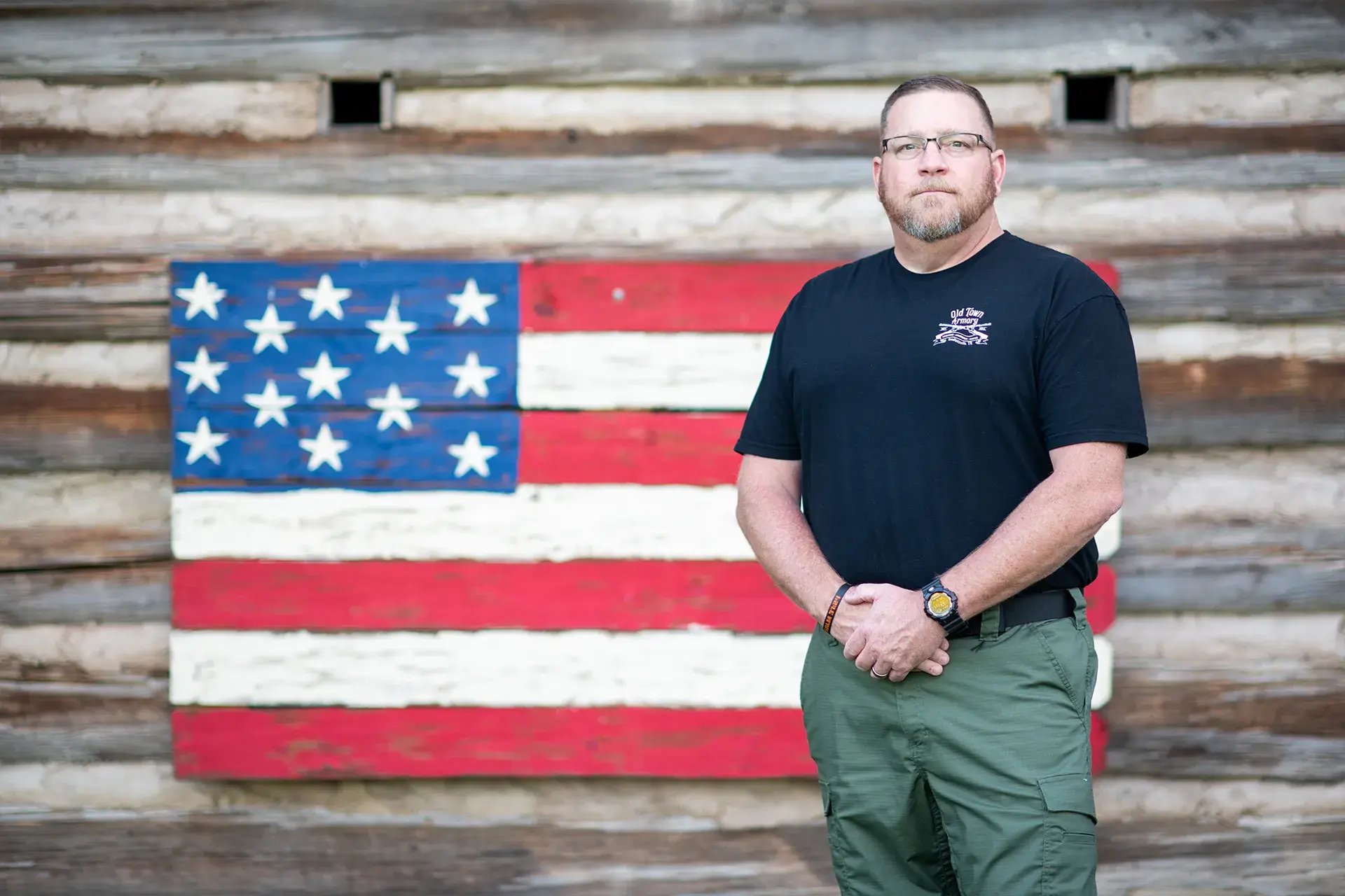 A man with short hair and glasses stands in front of a wooden wall with a painted American flag. He wears a black t-shirt and green pants, and has a watch on his left wrist.