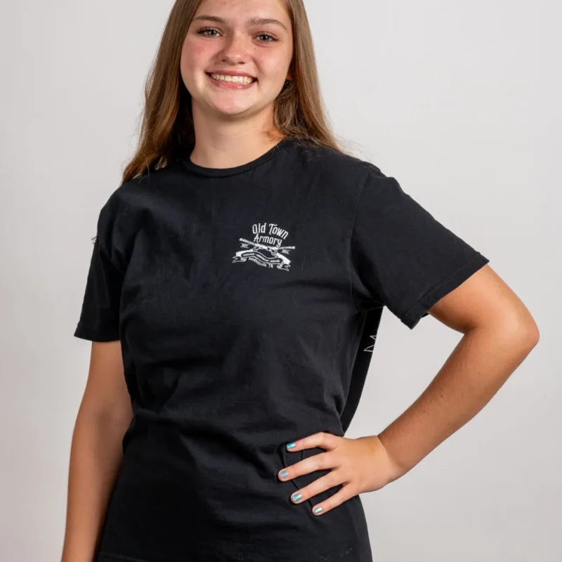 A person with long brown hair is smiling, standing with one hand on their hip, wearing a black "Old Town Archery" t-shirt against a plain white background.