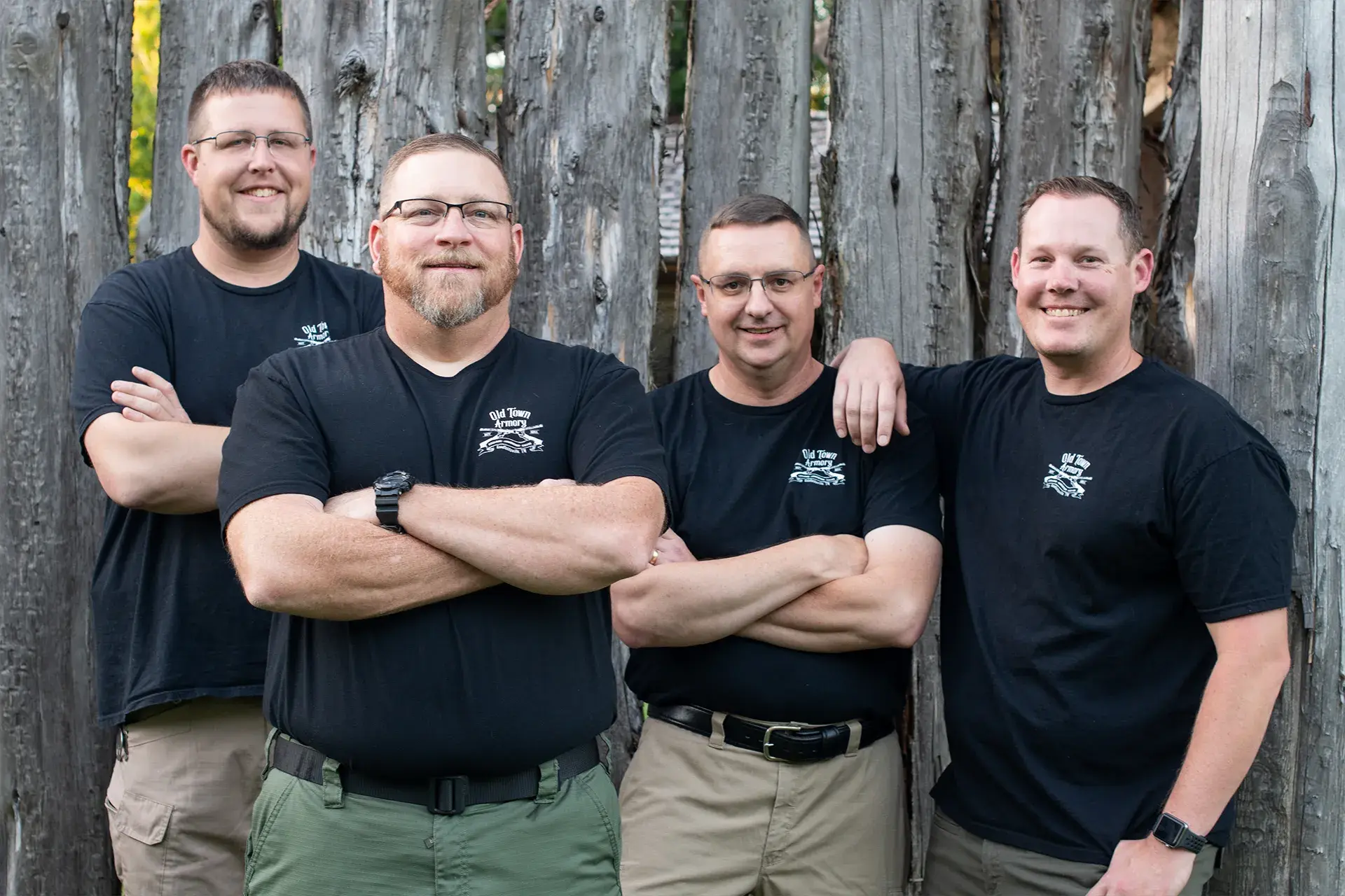 Four men in black shirts and khaki pants stand in front of a wooden fence, arms crossed or resting on one's shoulder, smiling at the camera.