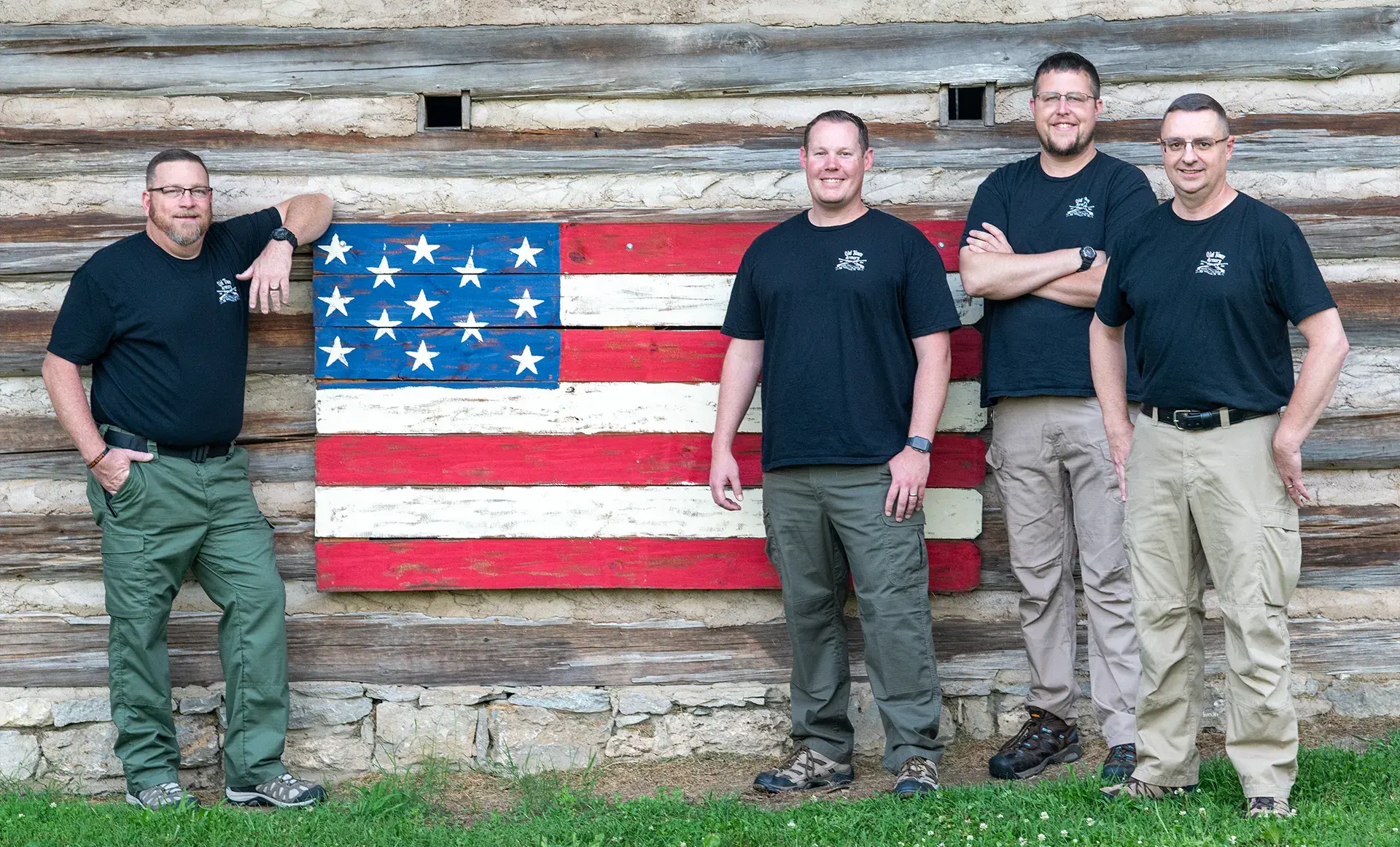 Four men in black shirts and khaki pants stand in front of a wooden wall featuring a painted American flag.