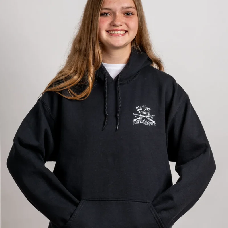 A person with long hair wearing a black hoodie with a white graphic on the left chest is standing against a plain background, smiling at the camera.