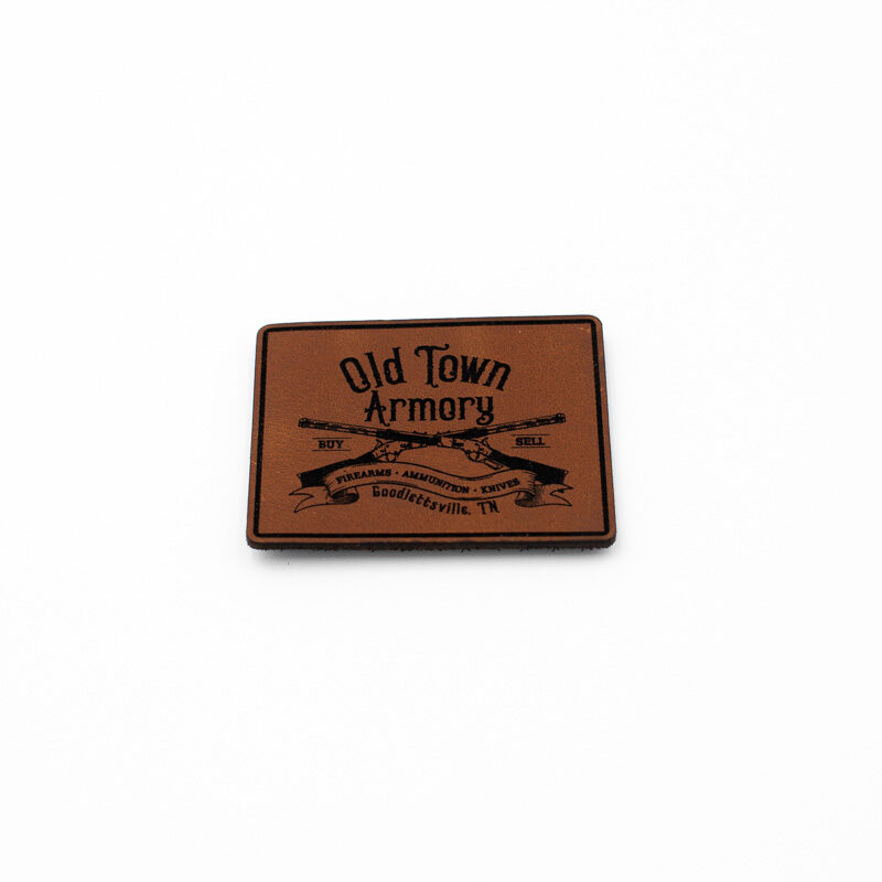 A brown square leather patch displaying the text "Old Town Armory" with crossed rifles graphic and additional smaller text.