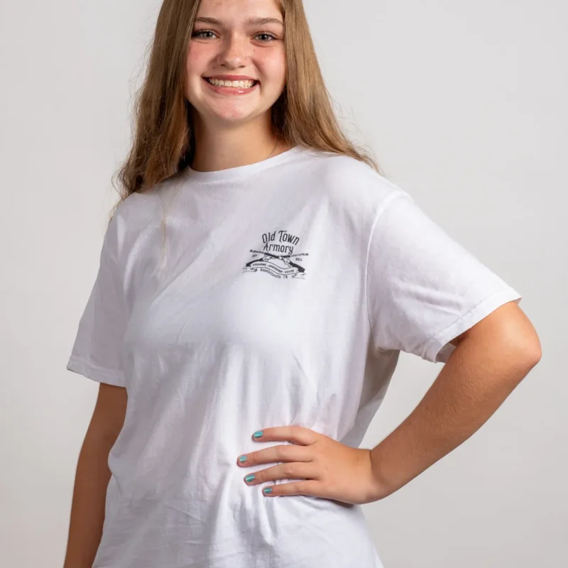 A young woman with long hair smiles and poses with her hand on her hip, wearing a white t-shirt with "Old Town Armory" printed on the front against a plain background.