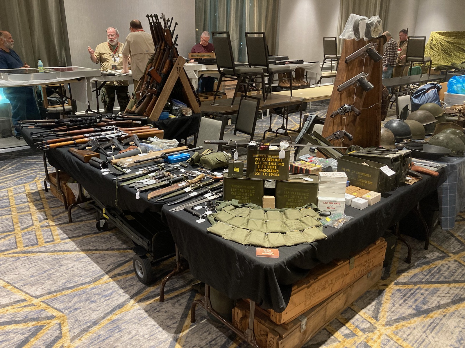 A display table with various firearms, ammunition boxes, and military gear including helmets and pouches at a convention or trade show. People are standing and inspecting items in the background.