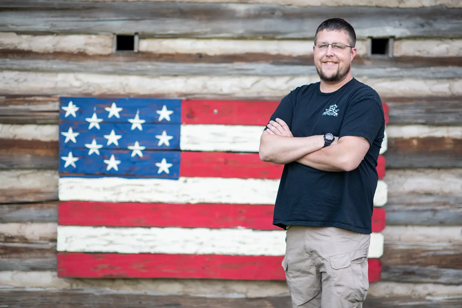 A man stands with arms crossed in front of a rustic wooden wall with a painted American flag, smiling at the camera.